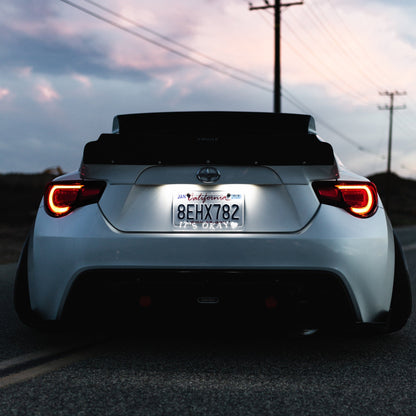Back view of a white car on a road with a clear license plate cover that says 'itsokay' with a sunset sky.