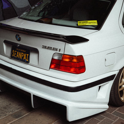 White BMW car with a black spoiler and California license plate and license plate cover that says 'itsokay' on a dark background