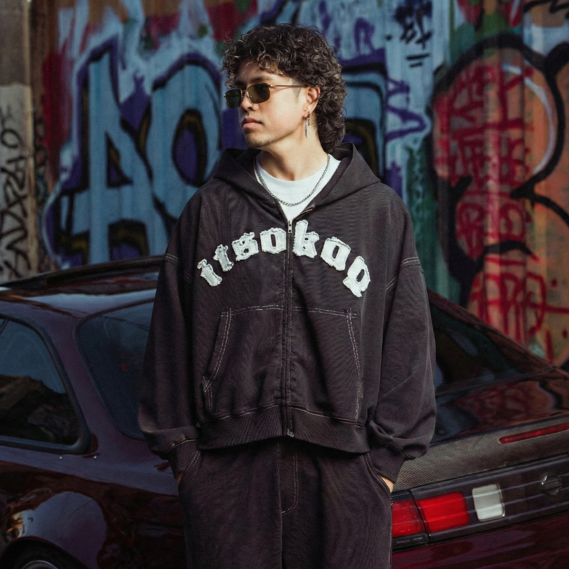 Person wearing a hoodie with text standing in front of a graffiti-covered wall and car.